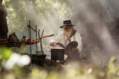 Kırsal bir kamp ateşinde yemek hazırlayan bir kovboy, yanında et pişirirken buharı tüten bir çorba tenceresini tatlandırıyor ve karıştırıyor.