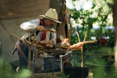 Kırsal bir kamp ateşinde yemek hazırlayan bir kovboy, yanında et pişirirken buharı tüten bir çorba tenceresini tatlandırıyor ve karıştırıyor.