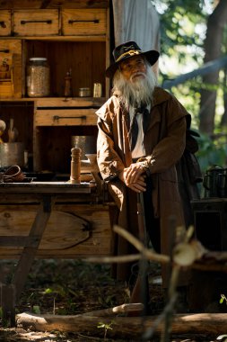 Orman kampında tüfeklere nişan alan kıdemli bir kovboy. Güneş ışığı dumandan yayılıyor ve arka planda bir yük arabası var.