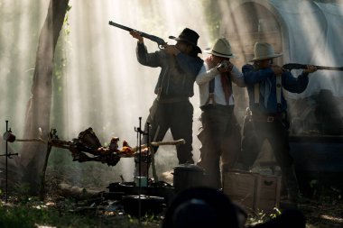 Bir orman kampında tüfeklere nişan alan kovboylardan oluşan bir grup. Arka planda da güneş ışığı bulutları ve bir yük arabası var.