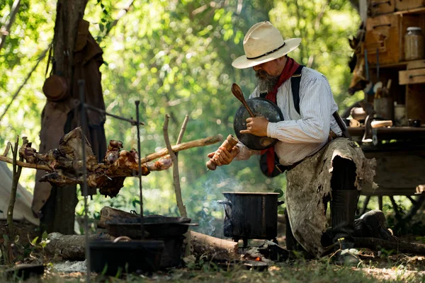 Kırsal bir kamp ateşinde yemek hazırlayan bir kovboy, yanında et pişirirken buharı tüten bir çorba tenceresini tatlandırıyor ve karıştırıyor.