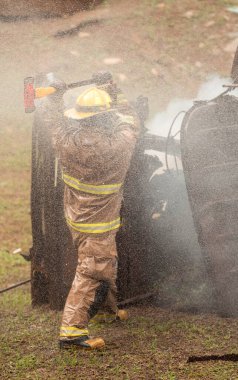 Tam donanımlı bir itfaiyeci canlı eğitim tatbikatı sırasında metal bir yapıya kurtarma baltasıyla vuruyor. Su püskürtme ve duman sahneyi doldurur.