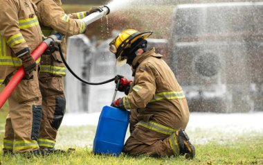 İtfaiyeciler canlı eğitim tatbikatı sırasında birlikte çalışırlar, yüksek basınçlı hortumlar kullanırlar bir ekip üyesi de destek ekipmanları hazırlar.
