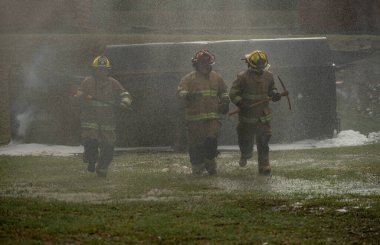 Bir grup itfaiyeci, acil durum tatbikatı sırasında ellerinde baltayla su buğusu ve ağır duman arasında koşuyor.