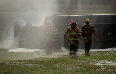 Bir grup itfaiyeci, acil durum tatbikatı sırasında ellerinde baltayla su buğusu ve ağır duman arasında koşuyor.