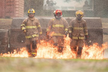 Tam koruma teçhizatı içinde bir grup itfaiyeci canlı bir yangın tatbikatı sırasında yerden alevler içinde yürür. Su püskürtme, duman ve yoğun ısı etraflarını sardı.