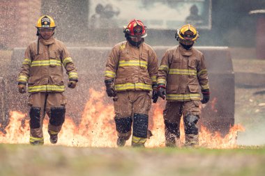 Tam koruma teçhizatı içinde bir grup itfaiyeci canlı bir yangın tatbikatı sırasında yerden alevler içinde yürür. Su püskürtme, duman ve yoğun ısı etraflarını sardı.