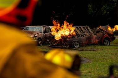 Yanan ve hasarlı araçlar yangın eğitimi senaryosu sırasında alevler içinde kalırken, itfaiyeciler olay yerini uzaktan izliyorlar.