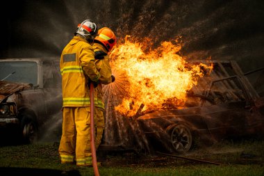 İki itfaiyeci güçlü bir su akışını büyük bir yangına doğru yönetirken pozisyonlarını aldılar. Yoğun ateş ve turuncu ışıltı koruyucu elbiseleriyle