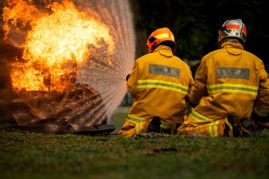 İki itfaiyeci güçlü bir su akışını büyük bir yangına doğru yönetirken diz çökerler. Yoğun ateş ve turuncu ışıltı koruyucu elbiseleriyle