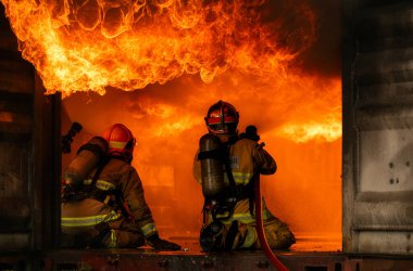 Tam koruyucu giysili iki itfaiyeci yoğun alevler ve dumanla çevrili büyük bir yangını söndürmek için birlikte çalışırlar.