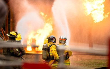 Bir grup itfaiyeci, yüksek basınçlı hortumlarla canlı bir yangın eğitimi sırasında büyük bir alevle savaşıyor. Ekip odaklanarak birlikte çalışırken parlak alevler sahneyi aydınlatıyor.