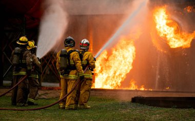 Bir grup itfaiyeci, yüksek basınçlı hortumlarla canlı bir yangın eğitimi sırasında büyük bir alevle savaşıyor. Ekip odaklanarak birlikte çalışırken parlak alevler sahneyi aydınlatıyor.