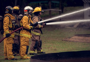 Bir grup itfaiyeci, yüksek basınçlı hortumlarla canlı bir yangın eğitimi sırasında büyük bir alevle savaşıyor. Ekip odaklanarak birlikte çalışırken parlak alevler sahneyi aydınlatıyor.