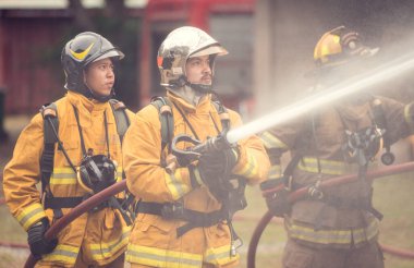 Bir grup itfaiyeci, yüksek basınçlı hortumlarla canlı bir yangın eğitimi sırasında büyük bir alevle savaşıyor. Ekip odaklanarak birlikte çalışırken parlak alevler sahneyi aydınlatıyor.