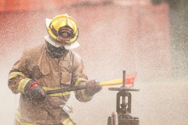 Koruyucu giysili bir itfaiyeci yangın musluğuna acil durum müdahalesi yaparken baltasını sallıyor. İtfaiyeci tehlikeli koşullar altında çalışırken su havaya sıçrıyor.