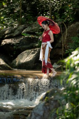 Geleneksel Tayland kıyafetleri içinde genç bir Asyalı kadın elinde kırmızı bir şemsiye tutarken yemyeşil bir tropikal ormanda küçük bir şelalede yürüyordu.