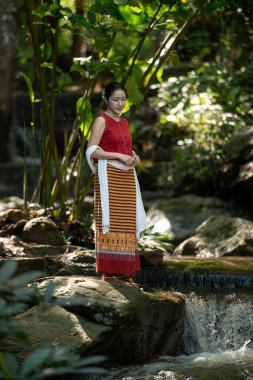 Geleneksel Tayland kıyafetleri içinde genç bir Asyalı kadın elinde kırmızı bir şemsiye tutarken yemyeşil bir tropikal ormanda küçük bir şelalede yürüyordu.