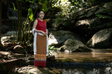 Geleneksel Tayland kıyafetleri içinde genç bir Asyalı kadın elinde kırmızı bir şemsiye tutarken yemyeşil bir tropikal ormanda küçük bir şelalede yürüyordu.