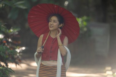 Doğayla çevrili bir bahçe yolunda yürürken elinde kırmızı bir şemsiye tutan geleneksel Tayland elbisesi giyen Asyalı kadın.