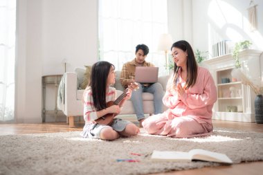 Babası arka planda dizüstü bilgisayarda çalışırken annesi için ukulele çalan genç bir kız.