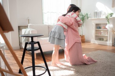 Bir anne yerde diz çöker ve kızına sıcak bir kucaklama yapar. Parlak, rahat bir oturma odasında, neşe ve güçlü aile bağlarını ifade eder.