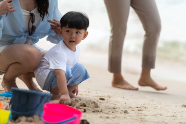 Tatilde, anne kumsalda arkasından yürürken çocuk kum oyuncaklarının yanında duruyor.