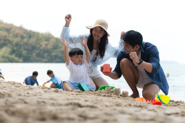 Tatilde, plajda birlikte oynayan genç bir aile, güneşli bir sahil tatili sırasında çocuklarının kumdan kale yapmalarına yardım eden aileler.