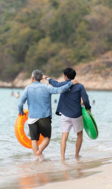 Tatilde, kıdemli adam ve oğlu gün içinde plajda şişme halkalar taşırken sığ deniz suyunda birlikte yürürler.