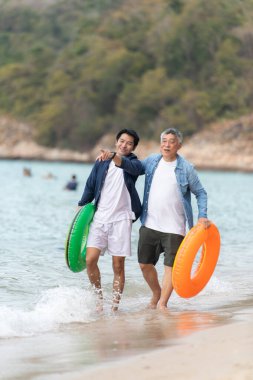 Tatilde, kıdemli adam ve oğlu gün içinde plajda şişme halkalar taşırken sığ deniz suyunda birlikte yürürler.