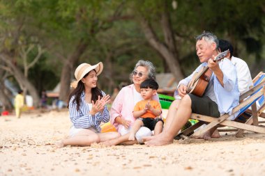 Yaşlı adam gitar çalarken, kum plajında birlikte oturan çok nesilli bir aile ve büyükanne ve çocuk gündüz vakti birlikte vakit geçiriyorlar.