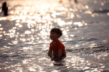Tatilde, küçük çocuk sığ deniz suyunda durup tek başına oynarken güneş ışığı gün batımında sahilde okyanusa yansıyor.