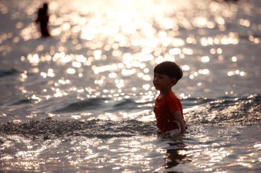 Tatilde, küçük çocuk sığ deniz suyunda durup tek başına oynarken güneş ışığı gün batımında sahilde okyanusa yansıyor.