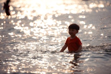 Tatilde, küçük çocuk sığ deniz suyunda durup tek başına oynarken güneş ışığı gün batımında sahilde okyanusa yansıyor.
