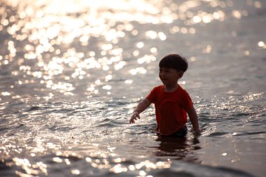 Tatilde, küçük çocuk sığ deniz suyunda durup tek başına oynarken güneş ışığı gün batımında sahilde okyanusa yansıyor.