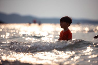 Tatilde, küçük çocuk sığ deniz suyunda durup tek başına oynarken güneş ışığı gün batımında sahilde okyanusa yansıyor.