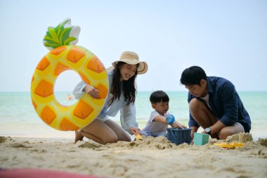 Tatilde ebeveynler, gün içinde deniz kenarındaki kumsalda birlikte oyuncak oynarken küçük çocuklarıyla birlikte duruyorlar.