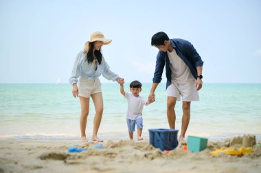 Tatilde ebeveynler, gün içinde deniz kenarındaki kumsalda birlikte oyuncak oynarken küçük çocuklarıyla birlikte duruyorlar.