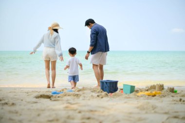 Tatilde ebeveynler, gün içinde deniz kenarındaki kumsalda birlikte oyuncak oynarken küçük çocuklarıyla birlikte duruyorlar.