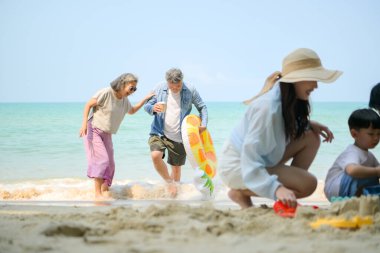 Tatilde ebeveynler çömelmiş ve küçük çocuklarıyla kum oyuncakları kullanarak oynarken yaşlı çift gün içinde deniz kenarında dinleniyor.
