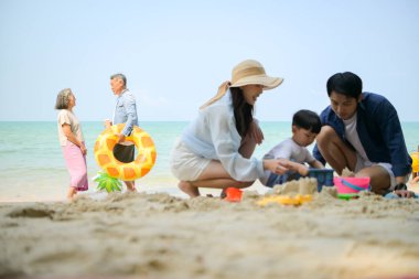 Tatilde ebeveynler çömelmiş ve küçük çocuklarıyla kum oyuncakları kullanarak oynarken yaşlı çift gün içinde deniz kenarında dinleniyor.