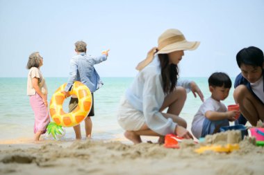 Tatilde ebeveynler çömelmiş ve küçük çocuklarıyla kum oyuncakları kullanarak oynarken yaşlı çift gün içinde deniz kenarında dinleniyor.