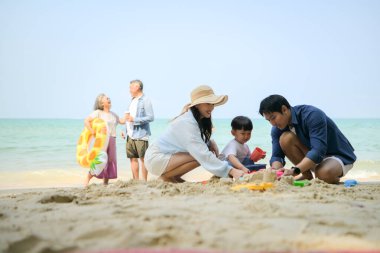 Tatilde ebeveynler çömelmiş ve küçük çocuklarıyla kum oyuncakları kullanarak oynarken yaşlı çift gün içinde deniz kenarında dinleniyor.