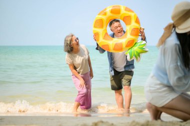 Tatilde ebeveynler çömelmiş ve küçük çocuklarıyla kum oyuncakları kullanarak oynarken yaşlı çift gün içinde deniz kenarında dinleniyor.