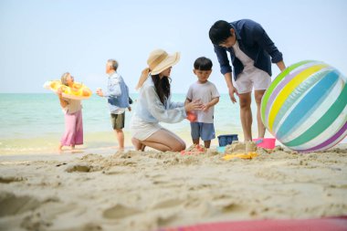 Tatilde ebeveynler çömelmiş ve küçük çocuklarıyla kum oyuncakları kullanarak oynarken yaşlı çift gün içinde deniz kenarında dinleniyor.
