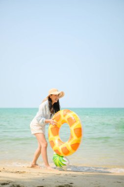 Tatilde, kumlu sahilde duran ve elinde şişme bir yüzük tutan genç bir kadın gün içinde deniz kenarında gülümserken.