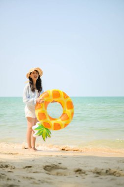 Tatilde, kumlu sahilde duran ve elinde şişme bir yüzük tutan genç bir kadın gün içinde deniz kenarında gülümserken.