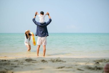 Tatilde, anne-babaların sığ deniz suyunda dikilip çocuklarını gün içinde okyanusa bakarken kaldırışlarının arka görüntüsü.