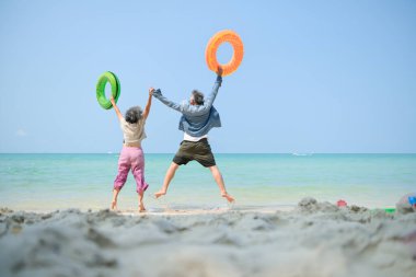 Tatilde, kıdemli çift sığ deniz suyunda durup gündüz vakti plajda şişme yüzük taşırken el ele tutuşuyorlar.