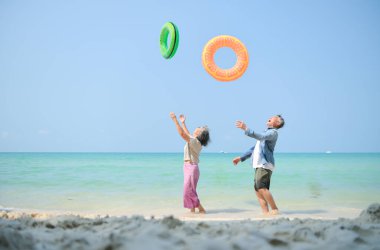 Tatilde, kıdemli çift sığ deniz suyunda durup gündüz vakti plajda şişme yüzük taşırken el ele tutuşuyorlar.